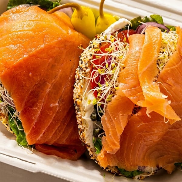 Two everything bagels with smoked salmon, cream cheese, sprouts, and vegetables in a takeout container.