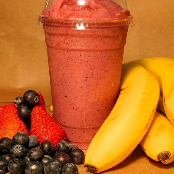 Berry smoothie in a clear cup surrounded by fresh strawberries, blueberries, and bananas.