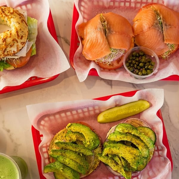 Assortment of bagel sandwiches with salmon, avocado, and cheese, served with pickles and fresh juices.